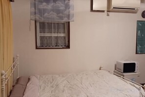 A bedroom with a bed, a window with a lace curtain, a bedside table with a microwave, and a wall-mounted air conditioner.