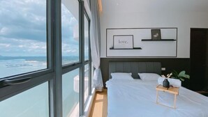 A modern hotel room with a large window, a bed with white linens, a bedside table with a vase and a plant, and a wall-mounted shelf with a framed picture and the word 'love.' - Seafront Country Garden Danga Bay by Lions Bay (Johor Bahru)