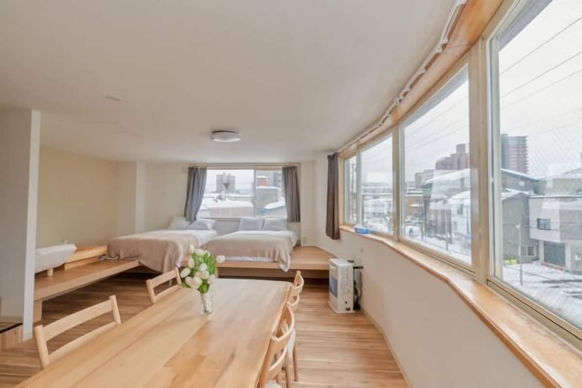 A modern hotel room with a wooden floor, a large window, a bed, a desk, and a small seating area.