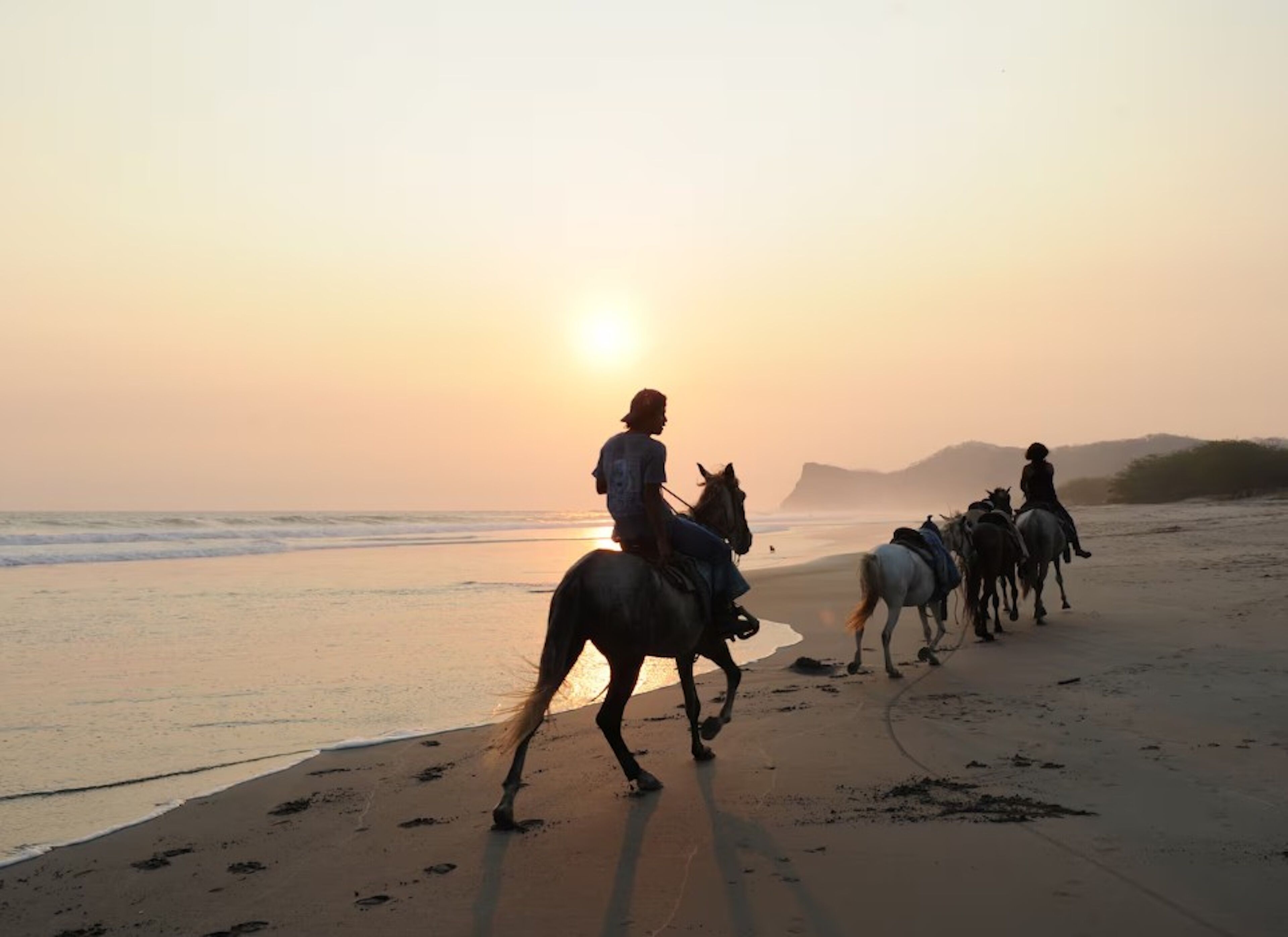 A beach scene with people riding horses along the shore during sunset.