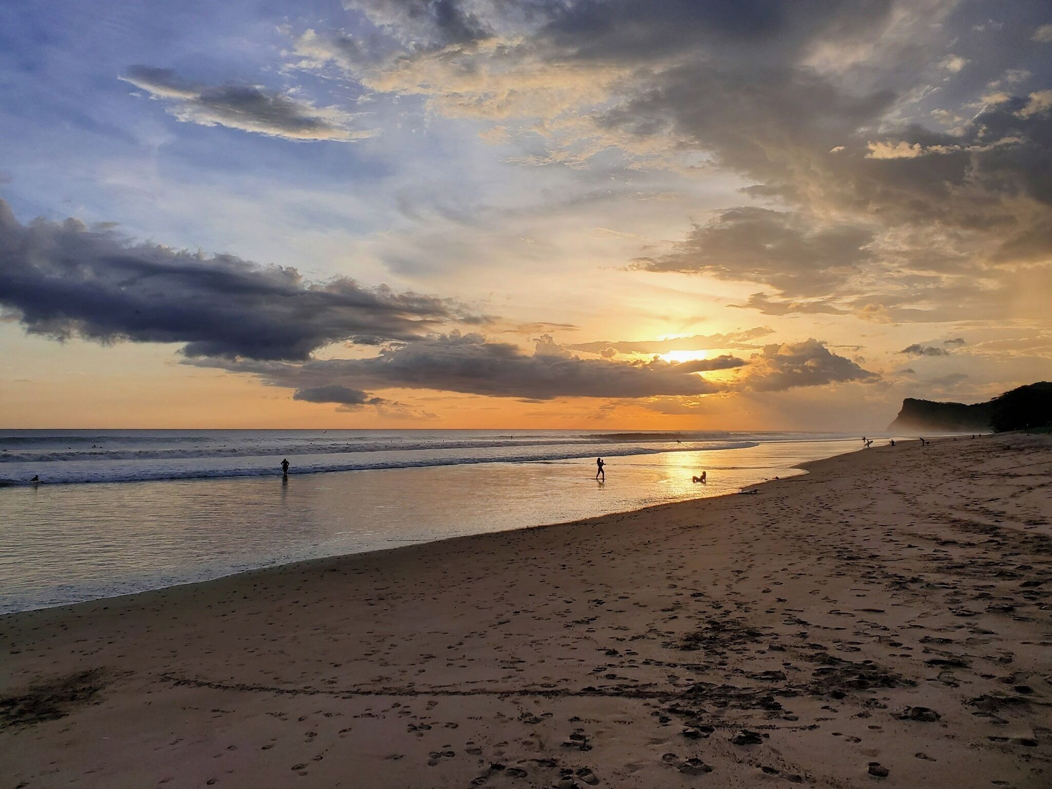 A beach with footprints and people walking, a sunset in the background, and a cliff in the distance.