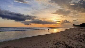 A beach with footprints and people walking, a sunset in the background, and a cliff in the distance.