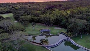 A golf course with a water hazard, surrounded by dense forest and a clear sky at sunset.