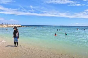A beach with clear water, people swimming, and a person standing on the shore. - Ocean Suites at One Pacific Residence - Mactan Newtown (Lapu-Lapu City)