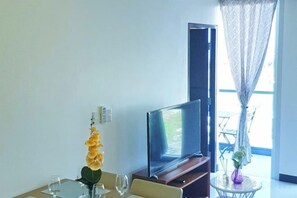 Een kamer met een flatscreen televisie op een standaard, een klein rond tafeltje met een vaas, en een raam met transparante gordijnen.