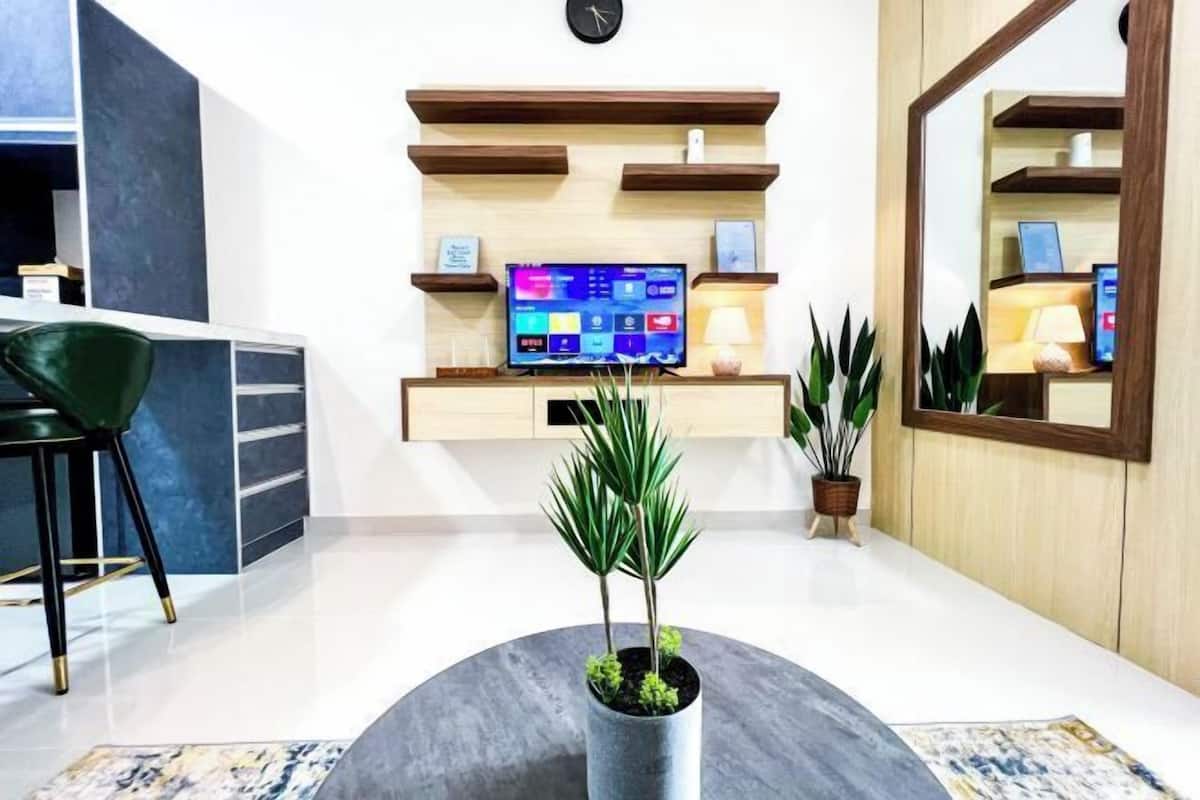 A modern living room with a flat-screen TV on a wooden shelf, a green chair, and a round coffee table with a plant.
