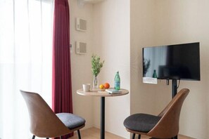 A room with two chairs, a small table with a plant, fruit, and a bottle, and a TV mounted on the wall. - KISETSU Kagurazaka (Tokyo)