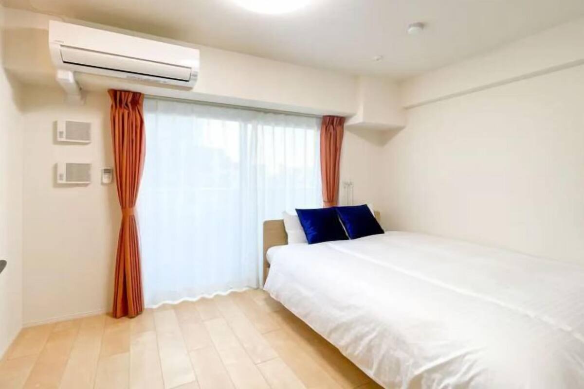 A hotel room with a bed, an air conditioning unit, a window with curtains, and a light switch on the wall.