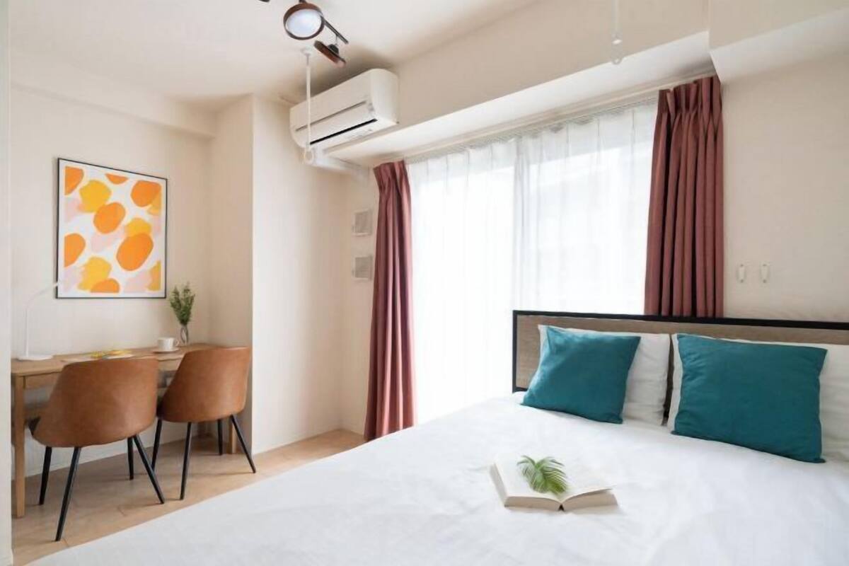 A hotel room with a bed, a desk, two chairs, a window with curtains, an air conditioner, and a wall-mounted artwork.