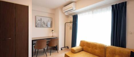 A compact living space with a sofa, a desk, and chairs, an air conditioning unit, and a window with curtains.