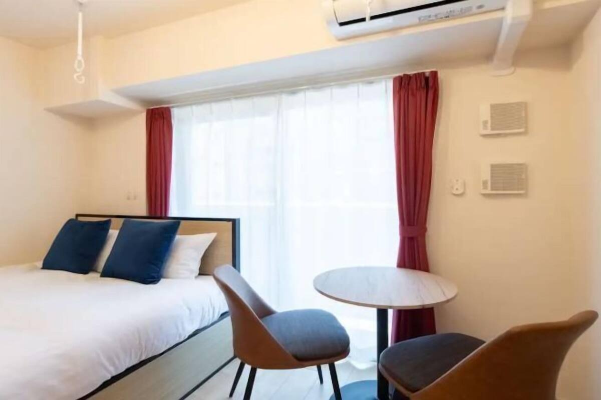 A hotel room with a bed, two chairs, a small table, and an air conditioning unit.