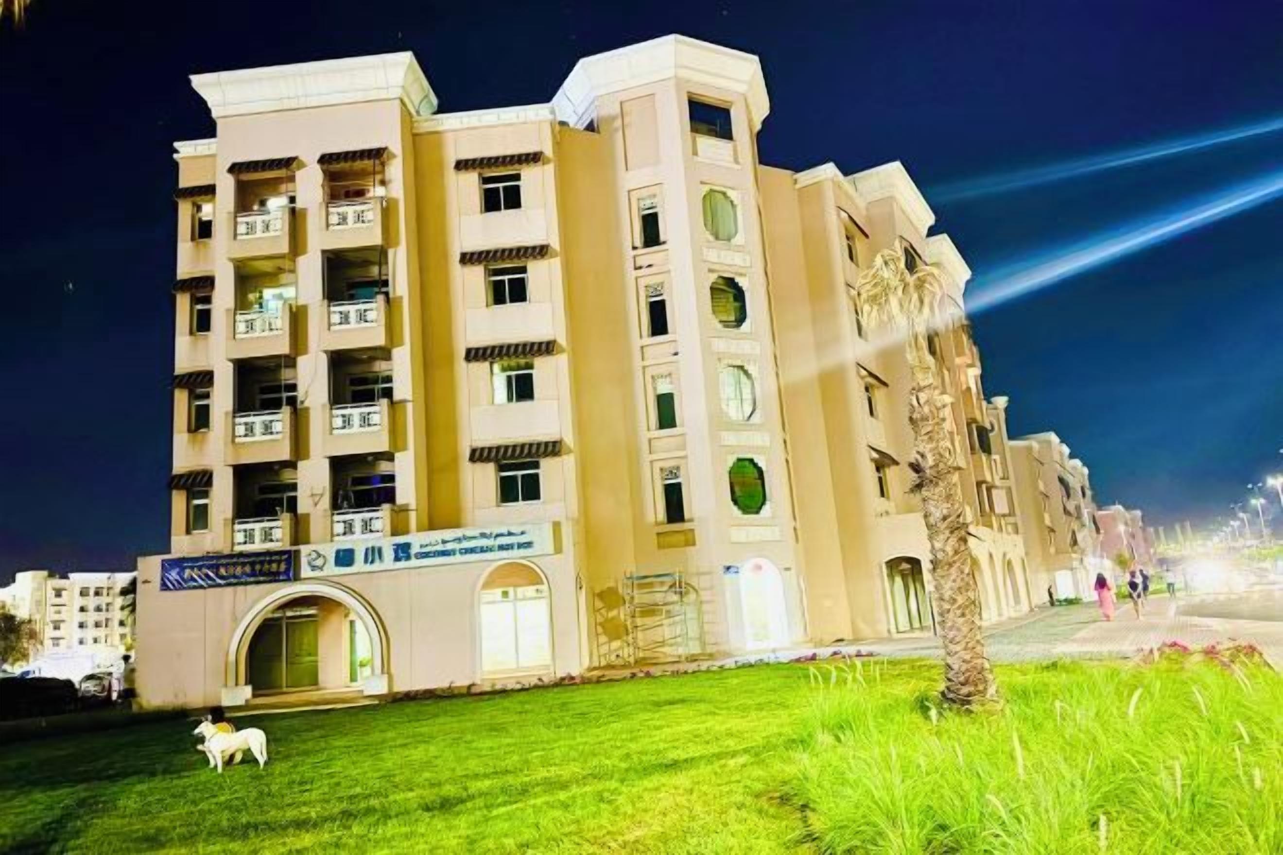 A multi-story building with balconies, a central entrance, and a grassy area with a white sculpture.