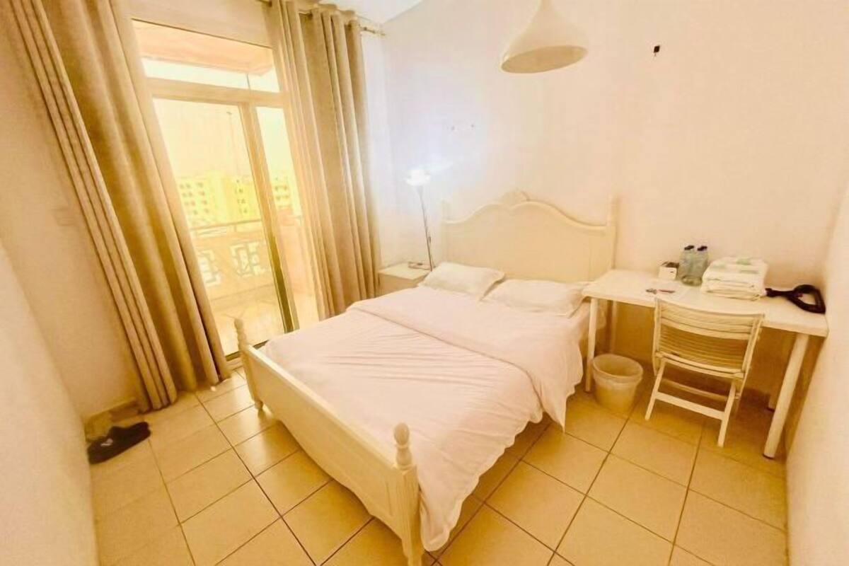 Une chambre d’hôtel avec un lit, un bureau, une chaise, une lampe et une vue sur les bâtiments environnants.