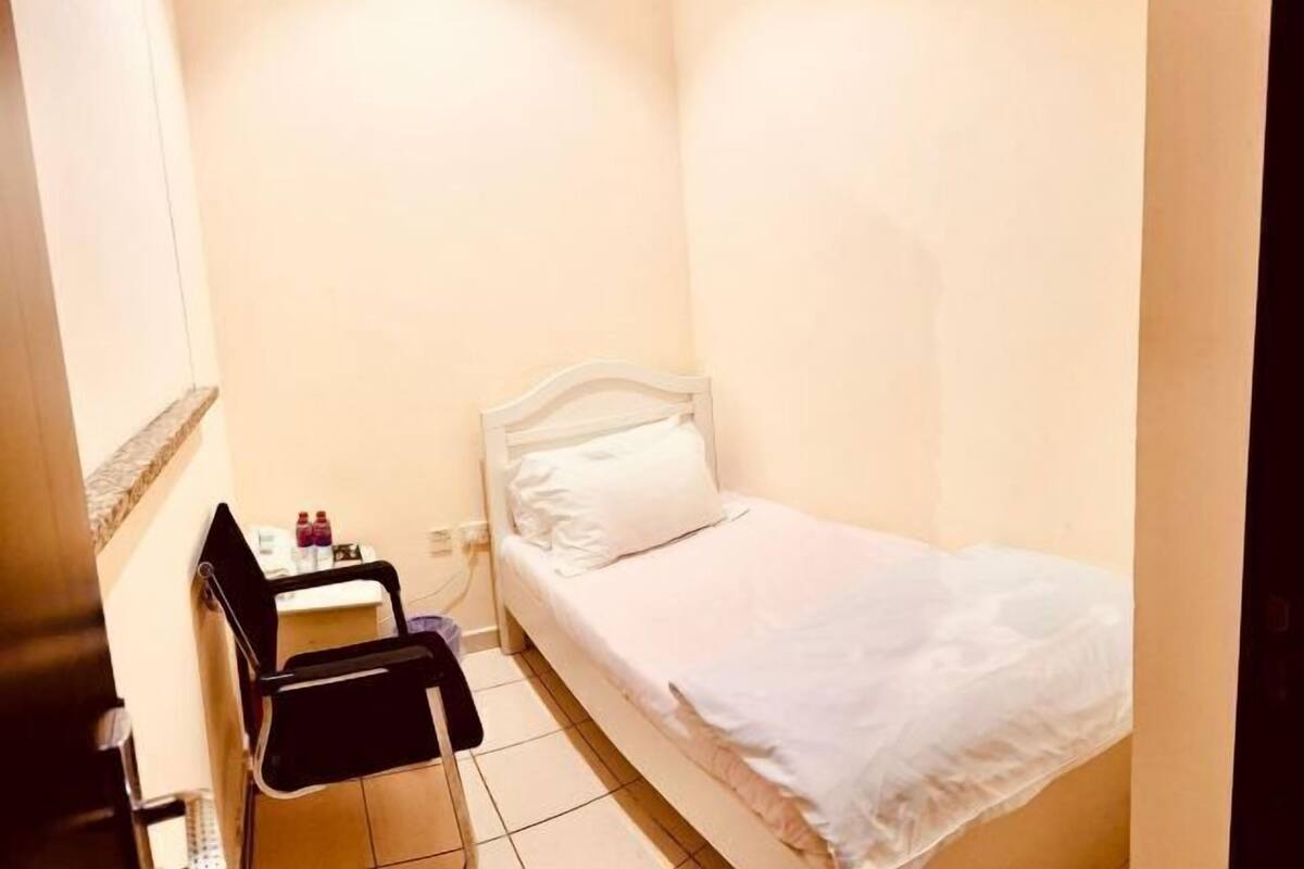 Une petite chambre avec un lit simple, une chaise et une table, une couvre-lit blanche et un mur uni.