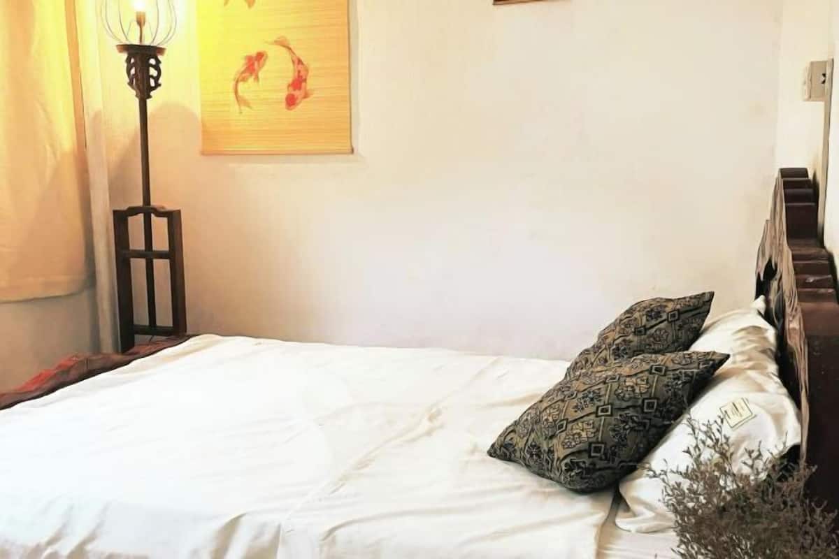 Un lit avec une literie blanche, deux coussins à motifs, une lampe sur pied et un mur orné d’un tableau encadré.