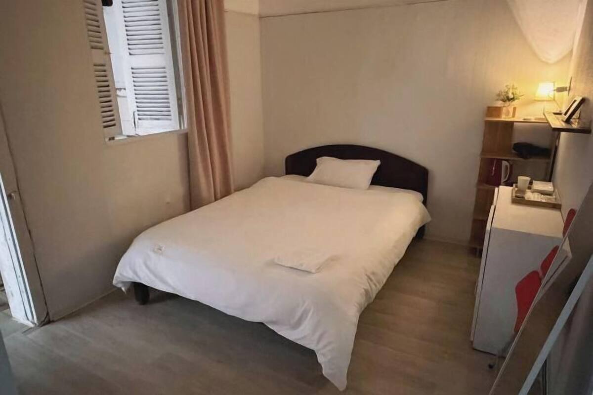 Un petit lit bien fait, avec une couette et des oreillers blancs, une table de chevet avec une lampe et une fenêtre dont les stores sont fermés.