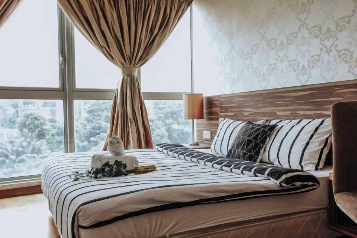 Una habitación de hotel con un ventanal grande, una cama con ropa de cama a rayas, un cabecero de madera y una mesita de noche con una lámpara.