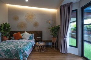 Un dormitorio moderno con cabecera de madera, cama con colcha estampada de motivos tropicales, mesita de noche con bandeja y un amplio ventanal con cortinas.