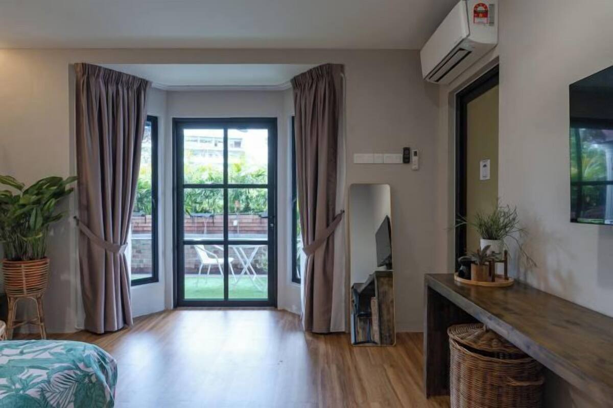 Una habitación con puerta corrediza de vidrio a un patio, una cama con estampado de hojas verdes, piso de madera, un televisor sobre un mueble bajo y un aire acondicionado instalado en la pared.