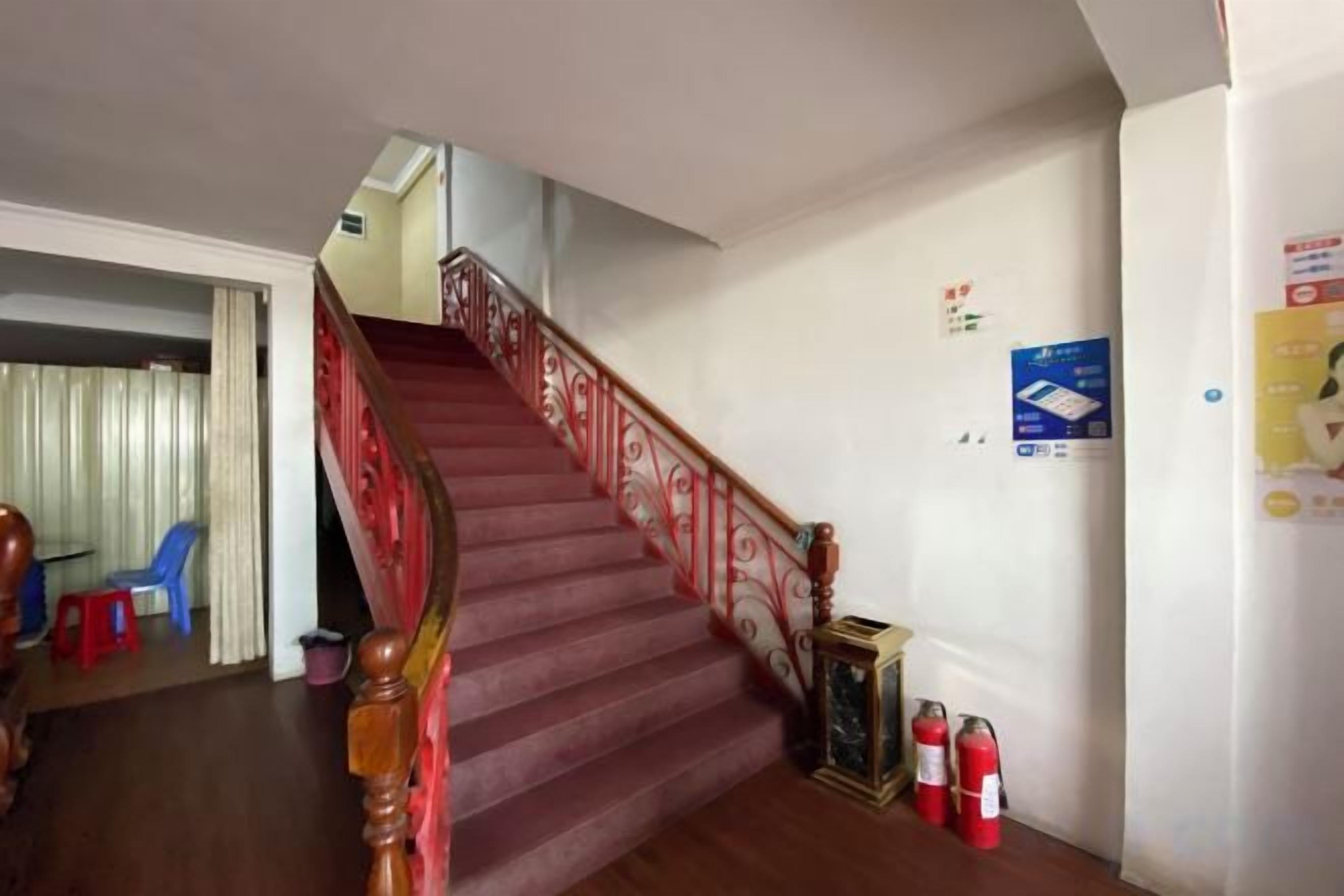 A staircase with a red handrail leading to an upper floor, a fire extinguisher on the wall, and a blue chair in the adjacent room.