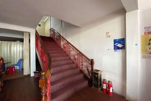 Un escalier avec une rampe rouge menant à l’étage supérieur, un extincteur fixé au mur et une chaise bleue dans la pièce adjacente.