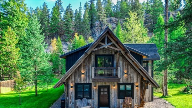 A wooden cabin with a balcony, a front porch with seating, and a gravel driveway. - The Solace by Bison Ridge Retreat (Whitefish)