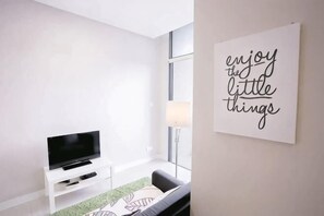 A modern living room with a flat-screen TV on a white stand, a green and white patterned rug, and a motivational quote on the wall.
