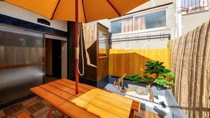 A wooden table under an umbrella, a small garden with a bonsai tree, and a building with a window. - AOU･SEITOKKU (Osaka)