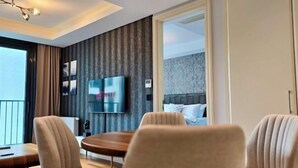 Una habitación de hotel moderna con un televisor de pantalla plana grande, una zona de comedor con mesa y sillas de madera, y vistas a la ciudad a través de amplios ventanales.