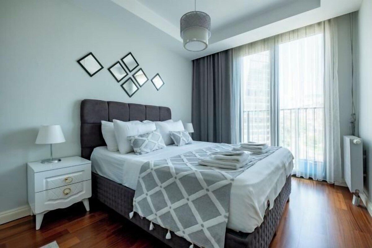 A modern bedroom with a large bed, a nightstand, a lamp, and a window with curtains.