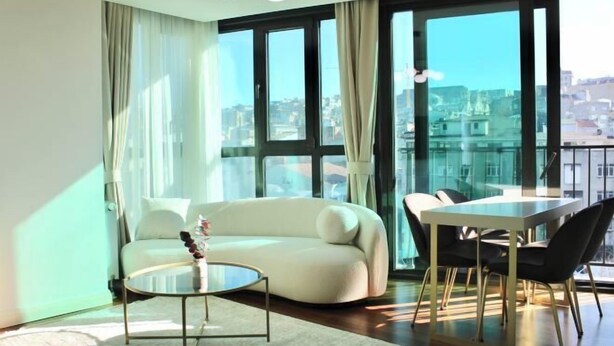 Una sala de estar moderna con un sofá, una mesita redonda, una mesa de comedor con sillas, ventanas amplias y vistas a la ciudad.