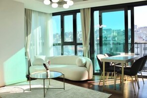 Een moderne woonkamer met een bank, een ronde salontafel, een eettafel met stoelen, grote ramen en een uitzicht op de stad.