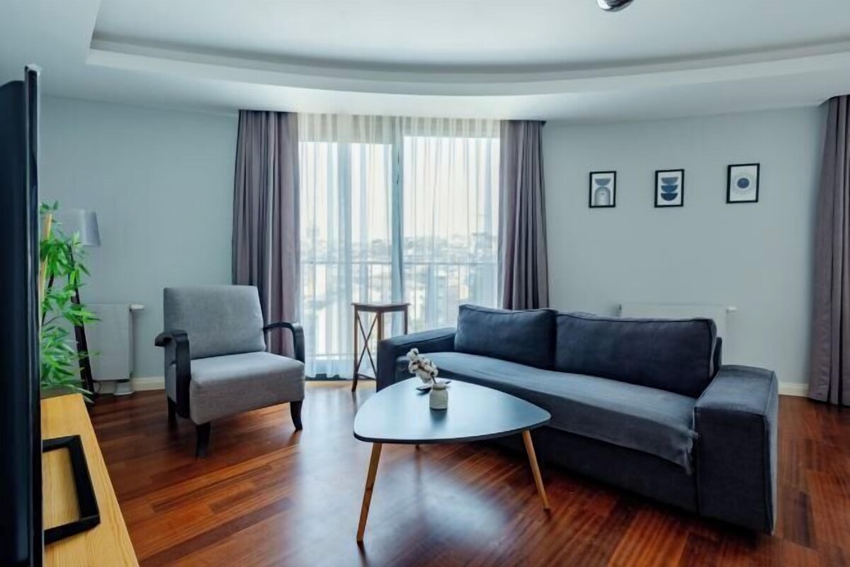 A modern living room with a grey sofa, a small round coffee table, and a grey armchair. There is a plant on a wooden table, and a window with curtains.