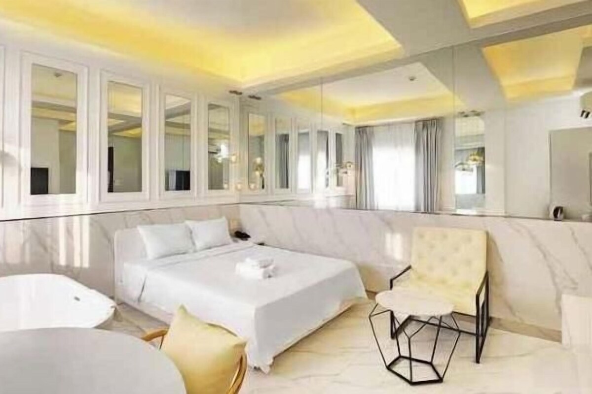 A modern hotel room with a large bed, a sitting area with a chair, a bathtub, and a large mirror.