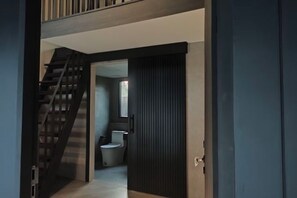 A modern interior with a staircase leading to a second floor, a dark sliding door, and a visible bathroom with a toilet and sink. - Ojisan House (Chon Buri)