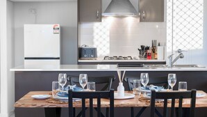 A modern kitchen with a central island, a built-in oven, and a gas stove. There is a dining table with chairs and a set of wine glasses. - Soothing 4-bedroom house with AC in Yarrabilba (Yarrabilba)