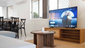 A modern living room with a TV displaying a show, a dining area with a table and chairs, and a cozy seating area with a sofa and a small coffee table. - Soothing 4-bedroom house with AC in Yarrabilba (Yarrabilba)