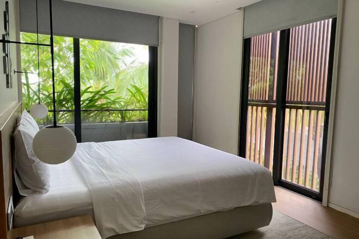 A modern bedroom with a large bed, a window with blinds, and a view of greenery outside.