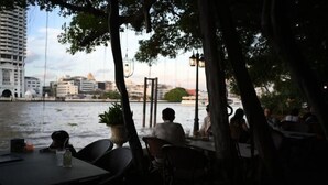 Un área de comedor al aire libre con mesas y sillas, con vista a un río y edificios al fondo.