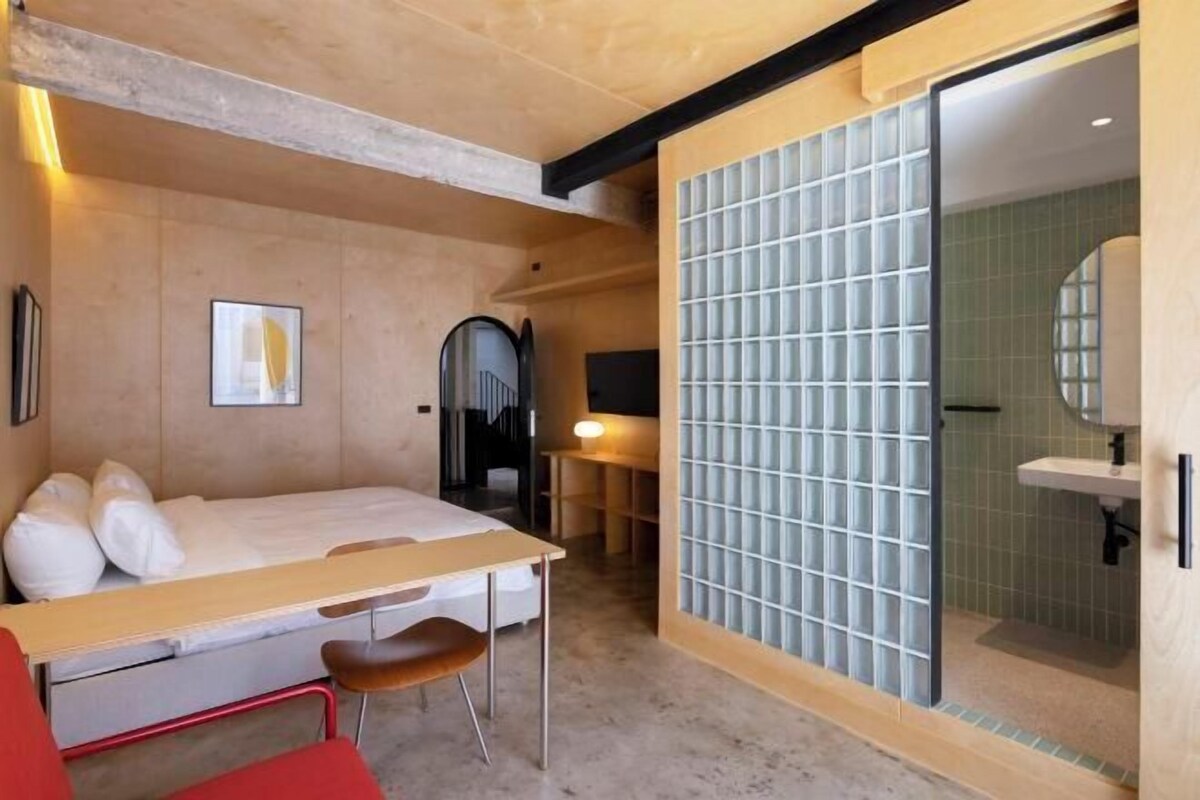 A bedroom with a bed, bedside table, chair, and a sliding glass door leading to a bathroom with a sink and mirror.