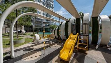 Un parque infantil con toboganes y estructuras de escalada, rodeado de un área con árboles y un edificio al fondo.