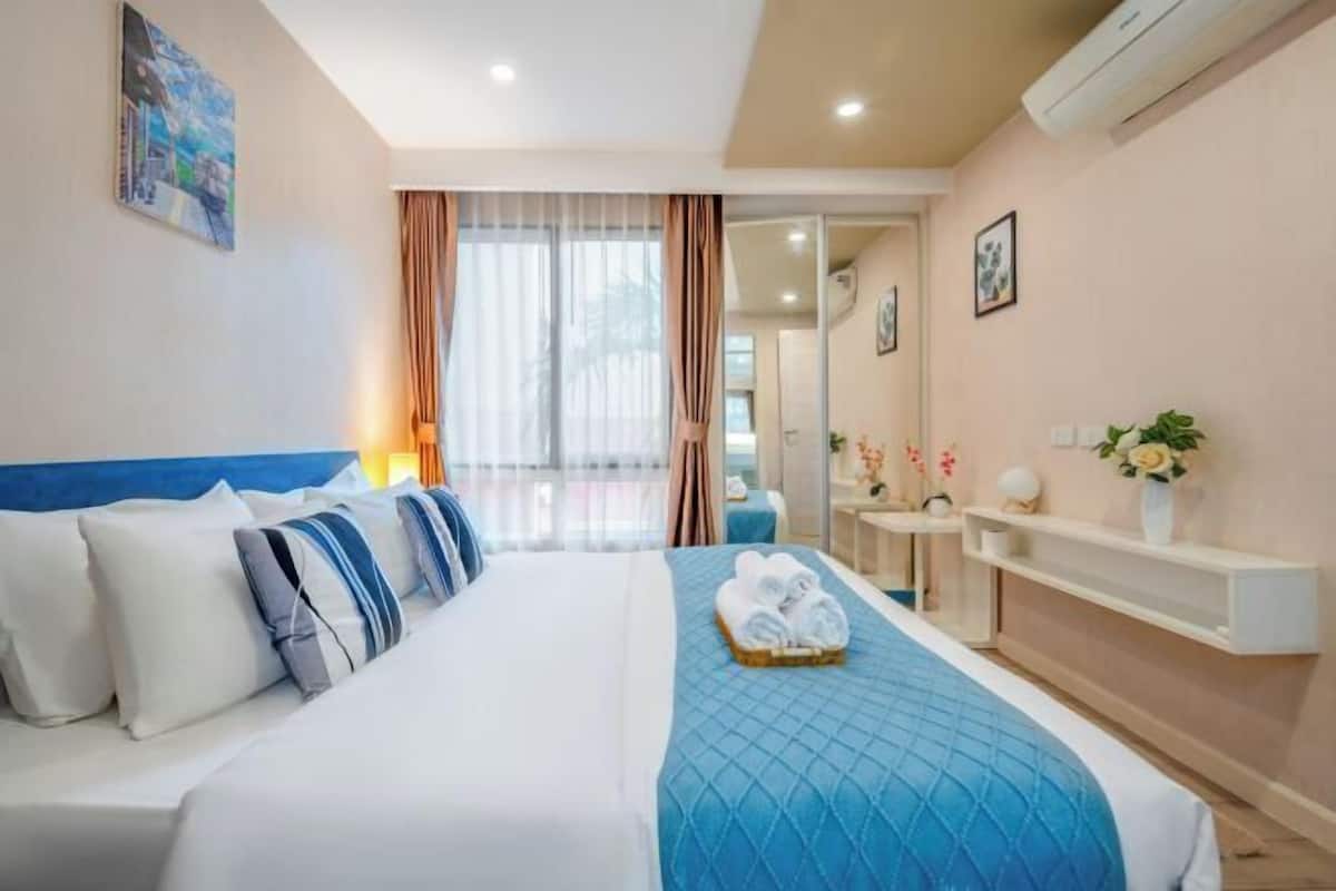 A hotel room with a bed, a bedside table, a mirror, a window with curtains, and a wall-mounted air conditioner.