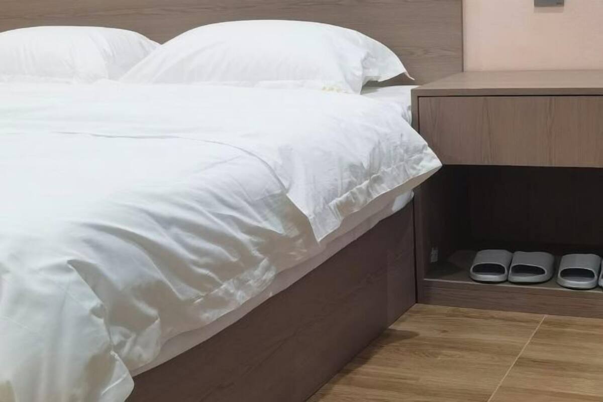 A bed with white bedding, a wooden headboard, and a nightstand with slippers.