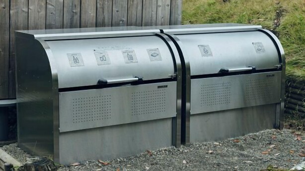 A metallic, curved trash bin with perforated sections and a lid, placed outdoors with a wooden fence in the background. - SANU 2nd Home Kawaguchiko1st (Narusawa)
