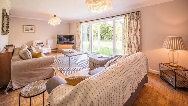 A modern living room with a sofa, armchair, and TV, a coffee table, and a large window with curtains.