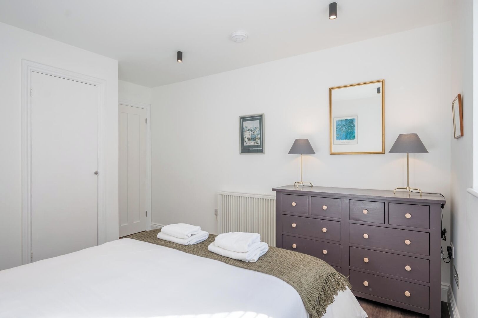 Een slaapkamer met een bed, een dressoir, twee lampen en twee prenten aan de muur.
