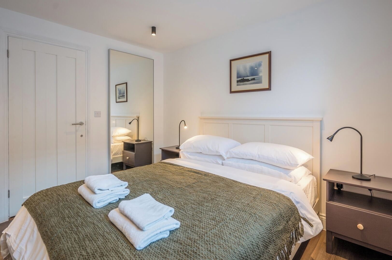 A neatly made bed with white linens and a green blanket, a bedside table with a lamp, a nightstand with a lamp, and a framed picture on the wall.