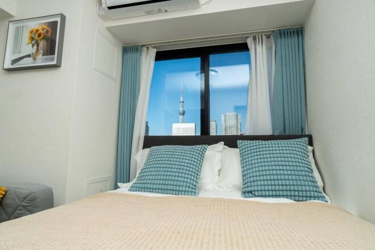 A hotel room with a bed, a window with curtains, a framed picture on the wall, and an air conditioning unit above the bed.