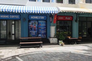 Un bâtiment avec un auvent bleu portant les inscriptions « HOANG COFFEE » et « HOMESTAY » sur le côté gauche, et un auvent rouge portant l’inscription « ADDI Hotel » sur le côté droit. Des bancs et des tables sont disposés à l’extérieur.