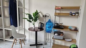 Une pièce de taille modeste, avec une chaise blanche, une petite table ronde ornée d’une plante, un distributeur d’eau et des étagères contenant divers objets.